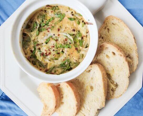 Overhead view of a small white ramekin of vegan spinach artichoke dip, next to some toasted slices of bread on a plate