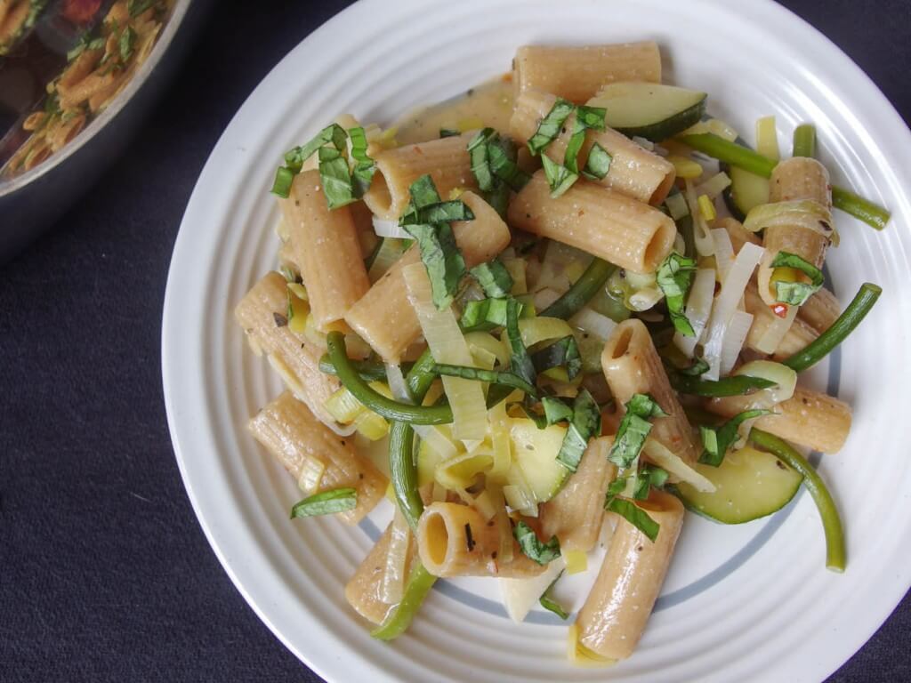 close-up of rigatoni pasta with zucchini, garlic scapes, and leeks served on a white plate garnished with basil