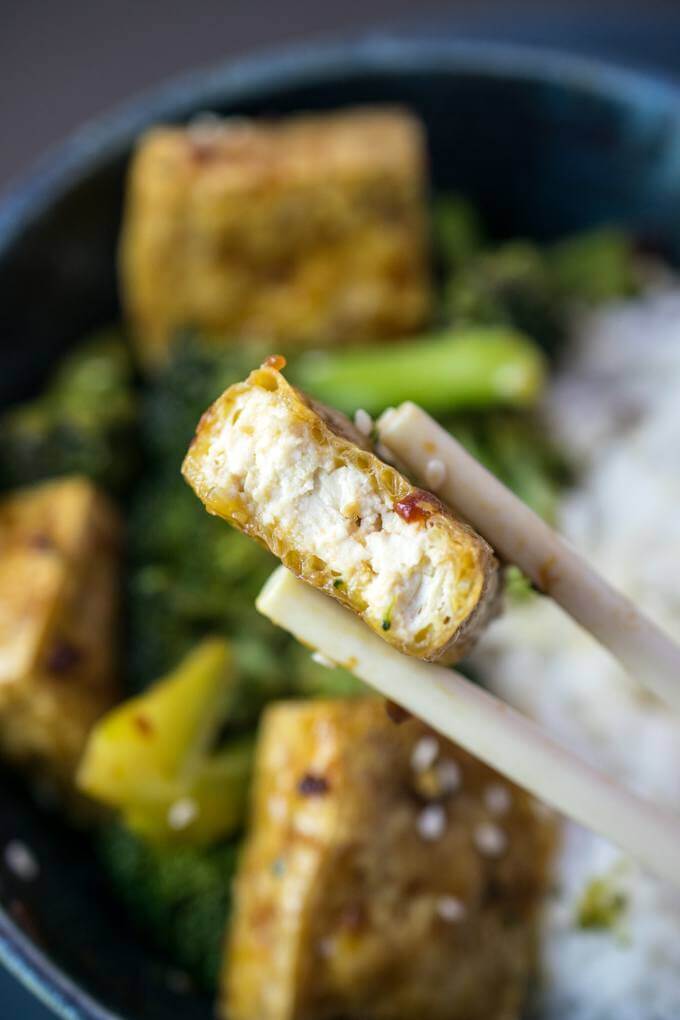 The inside of a cube of tofu bulgogi, showing a crispy exterior tofu skin drizzled with sauce, and a soft interior. A bowl of additional tofu and broccoli is in the background.