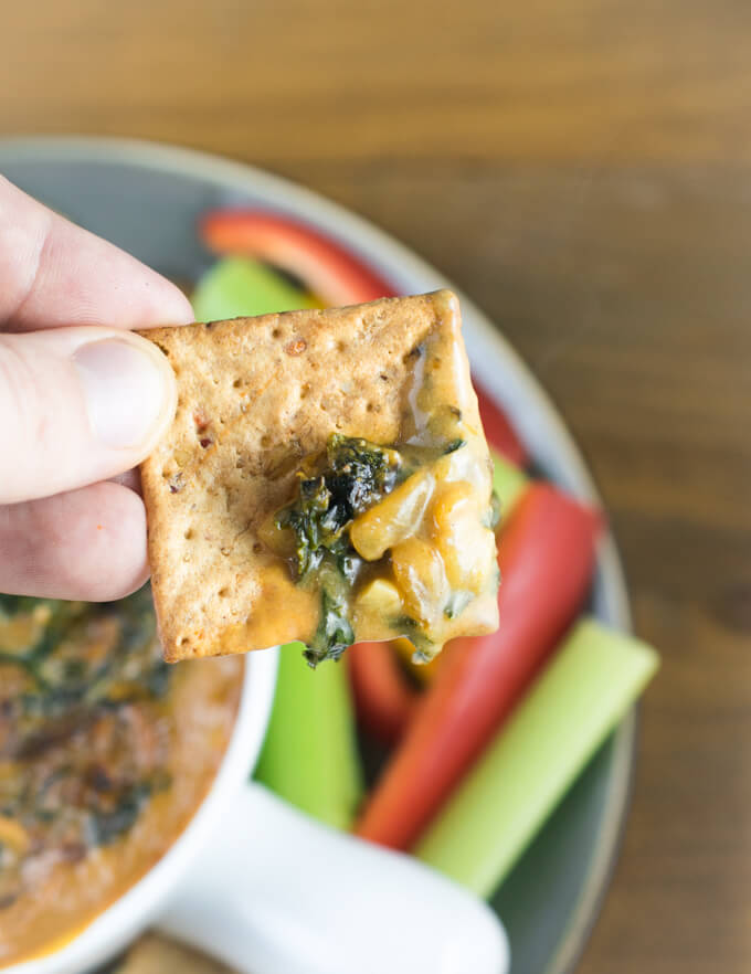 A little bit of vegan caramelized onion and kale dip spread onto a seedy cracker, showings it golden cheesy color and strands of wilted kale