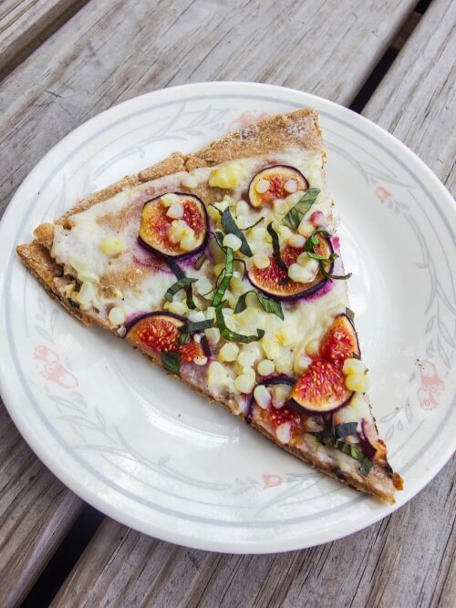 Close up of One slice of crispy vegan amaranth flatbread with sweet figs, corn, and fresh basil on a white floral plate outside on a picnic table