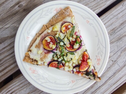 Slice of homemade flatbread pizza with corn, cashew cheese sauce, figs, and basil served on a floral plate