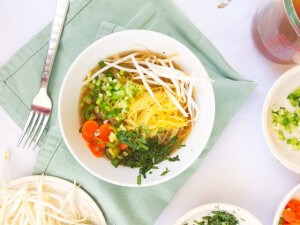 Assembled serving of real, nutrient filled Vietnamese noodle soup with hearty vegetable filling served on a blue kitchen towel with extra servings of crunchy bean sprouts and chopped green flavor