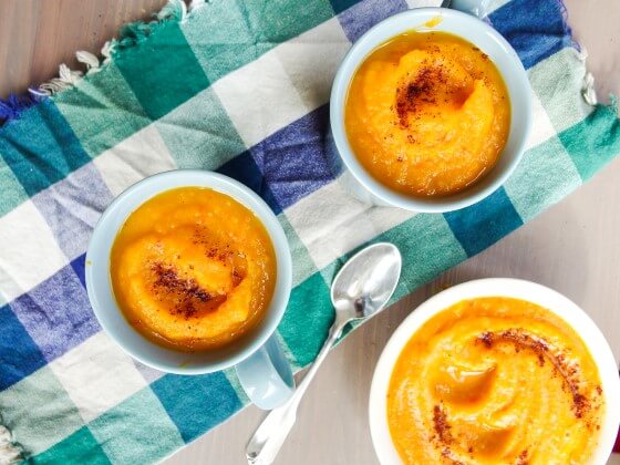 Three cups of light, nutrient dense acorn squash soup made with fresh whole foods and garnished with paprika. Served in white mugs on a blue checked kitchen towel