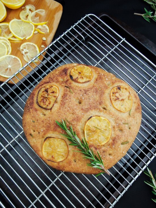 Closeup of a loaf of fluffy and light whole grain rosemary and meyer lemon foaccia made in the traditional dough sponge method served on a wire cooling rack with a fresh herb sprig | yupitsvegan.com