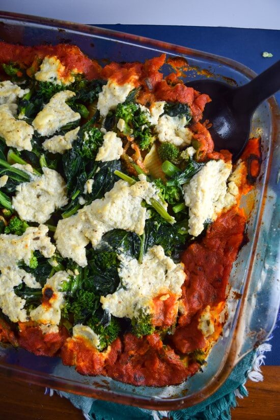 Close up of nut and refined sugar free baked pasta casserole with fresh green broccoli rabe, spicy arrabbiata pasta sauce, and cauliflower tofu ricotta in a baking dish