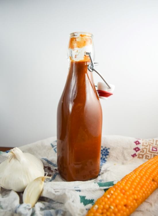 A flip top bottle of gluten and refined sugar free from scratch vegan barbecue sauce with intense umami and sweet flavors with a dash of whiskey - perfect comforting, healthy condiment for any meal. Served with corn!