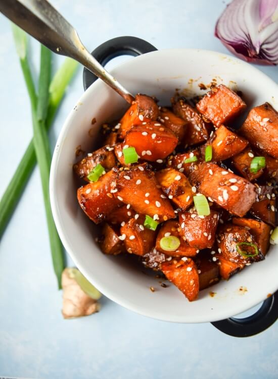 Oven roasted soft sweet potatoes served with a homemade teriyaki glaze. sliced green onions, spicy sriracha, and crunchy sesame seed in a white bowl