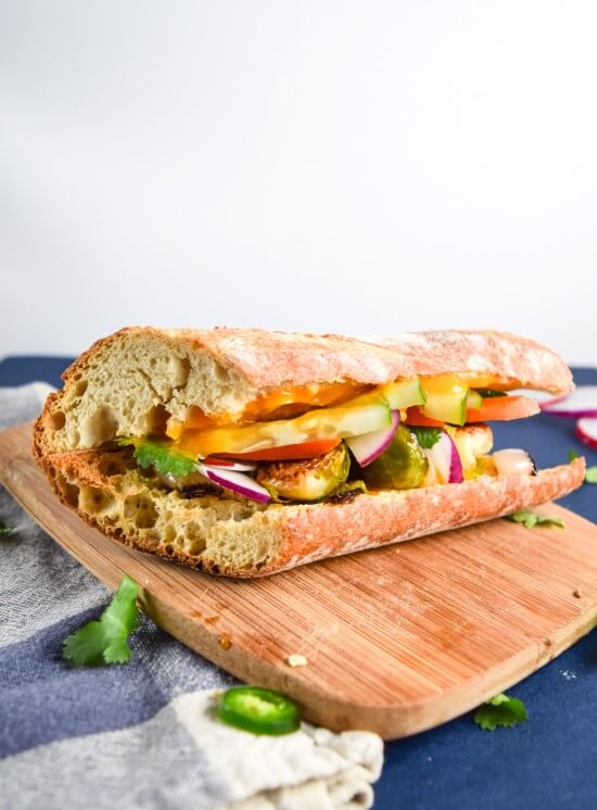 An assembled vegan bahn mi chay sandwich - spicy mango sauce, sweet oven roasted brussels sprouts and shallots, crunchy and crisp fresh vegetables, and fresh herbs on a fluffy crust French bread