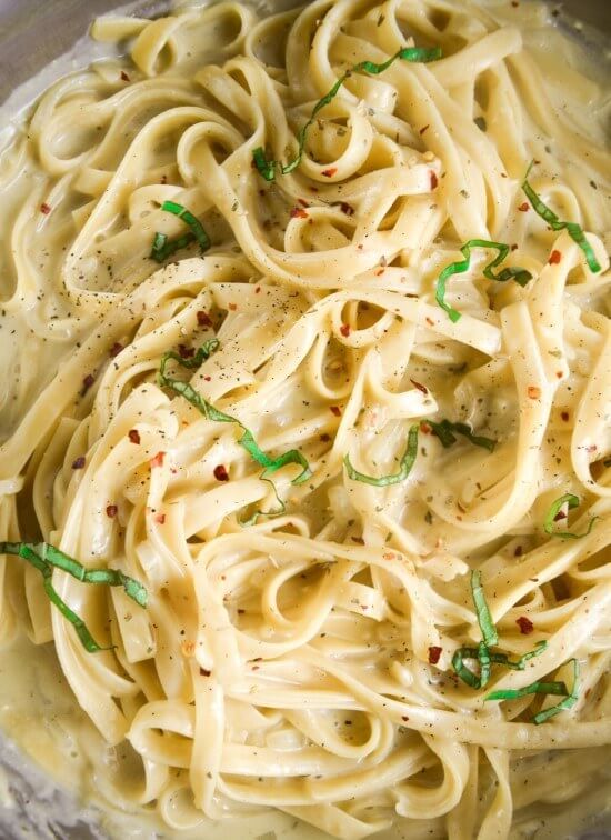 Close-up of creamy vegan fettuccine noodles in the pan, sprinkled with slivered basil, red pepper flakes, and ground black pepper.