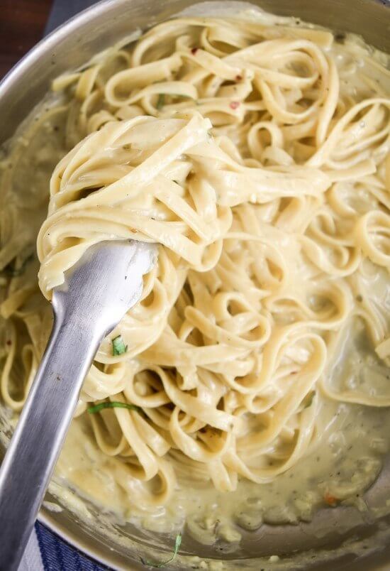 Vegan fettuccine alfredo being stirred in the pan using tongs; the sauce is visibly creamy with flecks of basil.