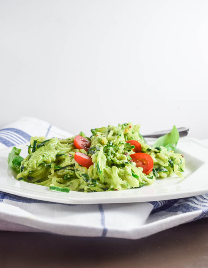 Luscious pesto inspired avocado and lemon juice sauce served over fresh cold zucchini noodles with juicy cherry tomatoes on a white plate