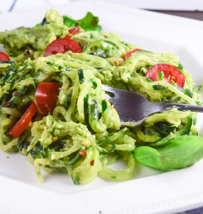 Close up of vegan zoodles with cucumber and zucchini, coated in rich avocado pesto sauce, garnished with red pepper and basil