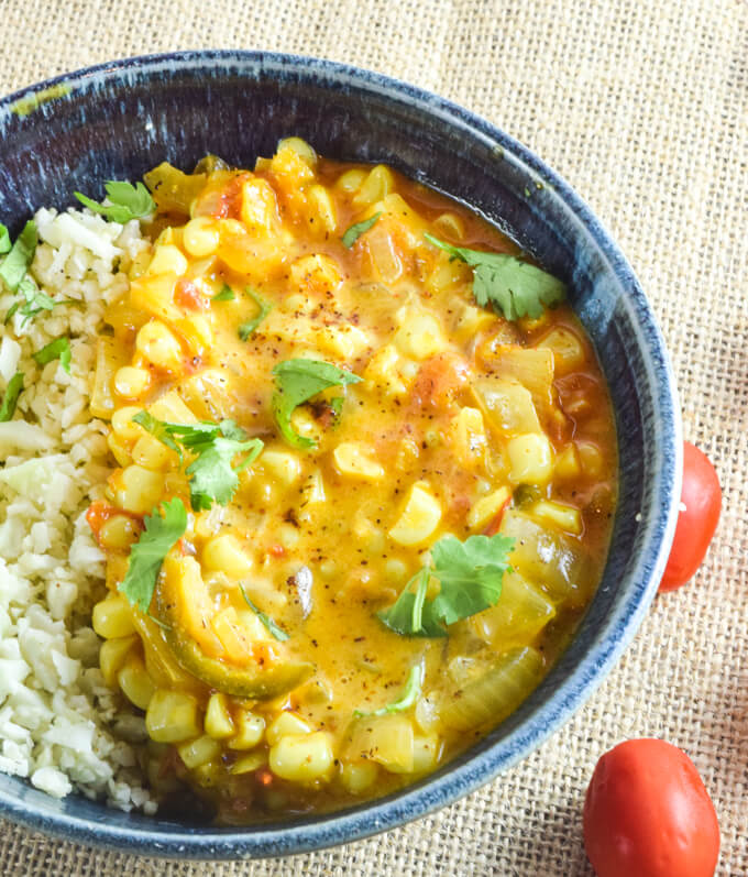 Close up of warming sweet corn curry with spicy peppers and onion, fresh tomatoes, creamy coconut milk and a cilantro garnish served with fluffy cauliflower rice in a blue bowl