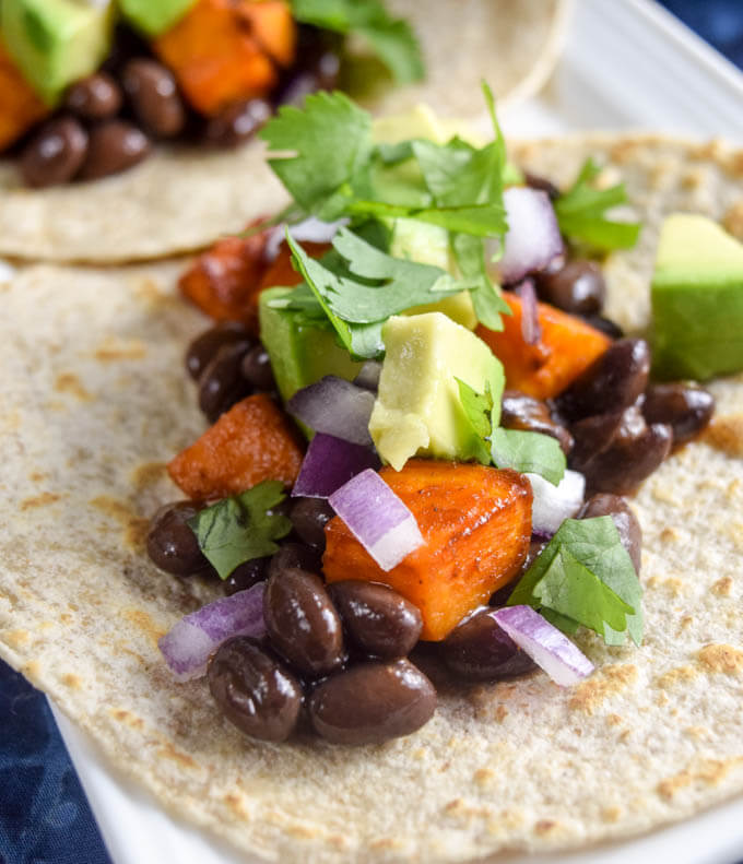 Closeup of bbq sweet potatoes, seasoned black beans, diced red onion, cilantro, and avocado in taco wraps