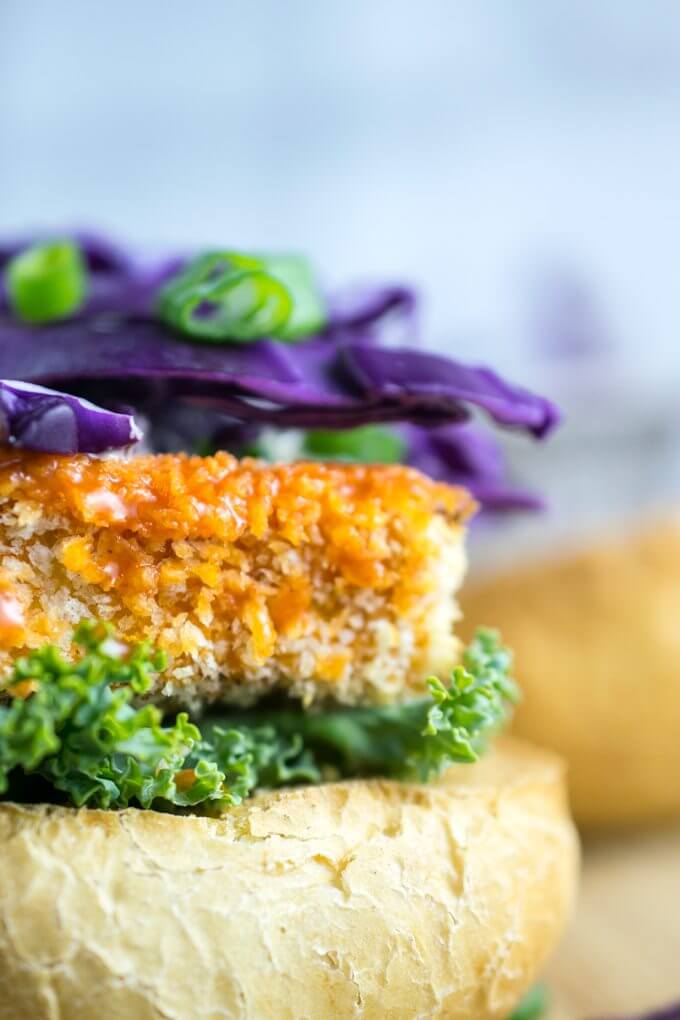 Closeup of buffalo tofu coated in crispy panko, topped with crunchy red cabbage, green onions, and fresh lettuce on a slide bun