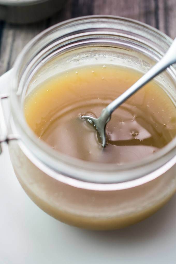 A glass jar of sweetened condensed macadamia nut milk on a white plate, with a spoon dipping into the jar showing the vegan condensed milk's thick and gooey texture.