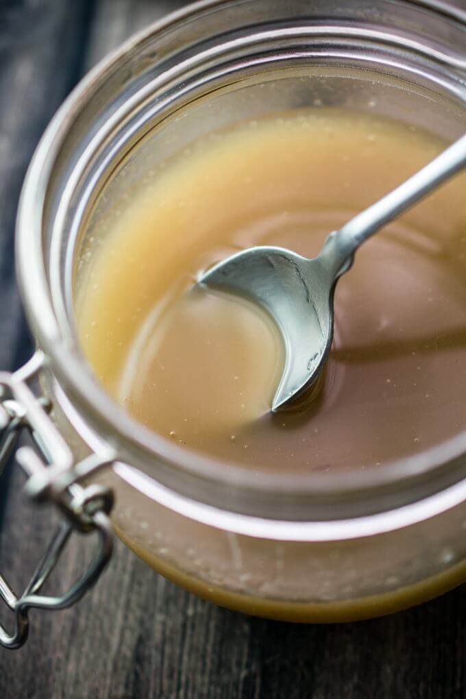 Vegan sweetened condensed milk in a glass jar on a gray wooden table, with a teaspoon being dipped into it.