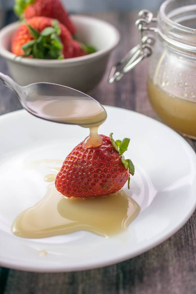 A fresh strawberry on a white plate, being drizzled with vegan sweetened condensed milk. In the background is a bowl of more strawberries and a jar of the paleo condensed milk.