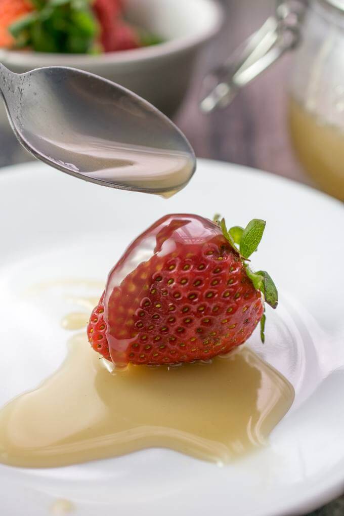 Sweetened condensed almond milk dripping off of a teaspoon onto a strawberry on a white porcelain plate.