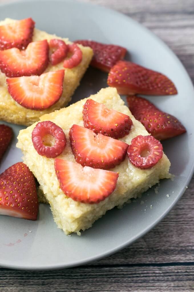 Two slices of tres leches cake arranged on a gray plate on a gray wooden table, drizzled with vegan macadamia nut syrup and topped with a layer of sliced strawberries and raspberries.