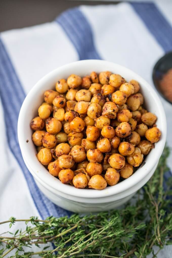Blackened chickpeas in a white bowl, garnished with fresh thyme, on a white and blue napkin next to a small bowl of Cajun seasoning.