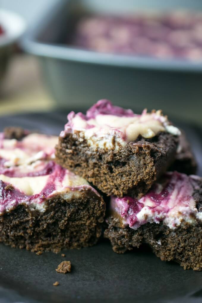 Vegan brownies stacked on a plate, with moist chocolatey interiors and topped with a white cashew cream and purple berry swirl.