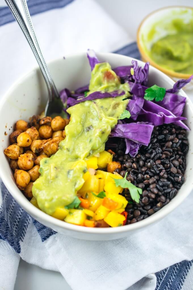 Breakfast burrito bowls with black rice, mango salsa, chopped purple cabbage, spiced chickpeas, and topped with avocado crema. An extra bowl of avocado cream is in the background.