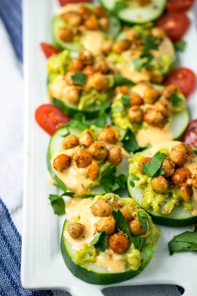 Cucumber appetizers lined up on a white plate with a blue and white napkin beneath it. Browned chickpeas and avocado top the cucumbers, with tomatoes in the background.