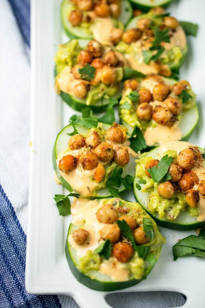 Cucumber rounds with mashed avocado and creamy chipotle sauce, topped with spiced chickpeas, lined up on a white plate and garnished with fresh parsley.