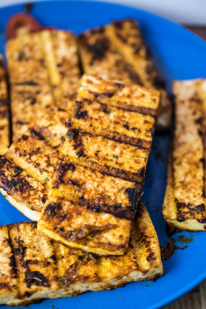 A blue plate stacked with pieces of grilled tofu gyros with charred grill marks, and cherry tomato slices in the background.