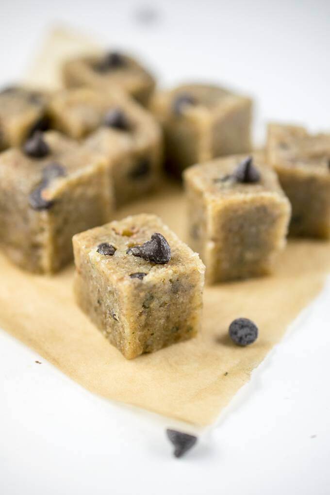 Sliced dairy-free cookie dough squares arranged on a white table with parchment paper, chocolate chips and hemp seeds.