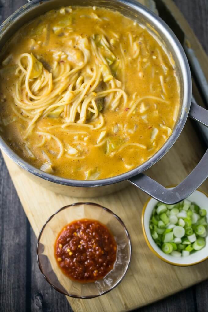 En stor kastrull med one pot spicy garlic almond butter noodles, arrangerad på en skärbräda bredvid en skål med chili-vitlökssås och en skål med schalottenlök.