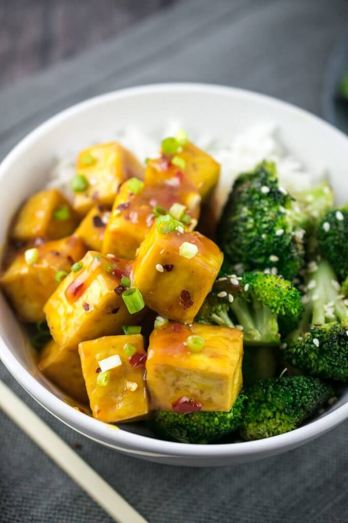 Baked orange tofu in a white bowl, garnished with green onions and red pepper flakes, next to stir-fried broccoli with sesame seeds
