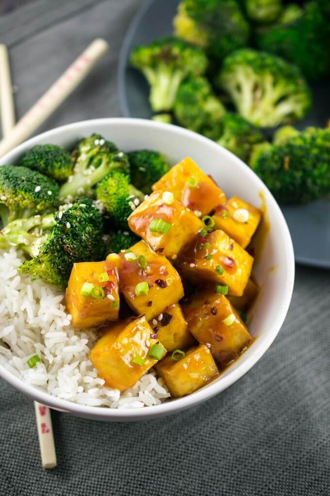 A bowl of orange-glazed tofu, white rice, and sesame broccoli with broccoli and chopsticks in the background