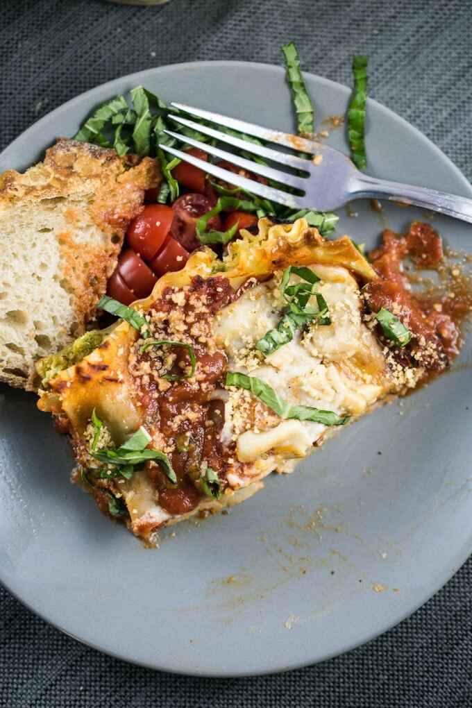 A slice of vegan lasagna on a gray plate on top of a dark gray napkin, with sliced cherry tomatoes, sourdough bread, and chiffonade of fresh basil.