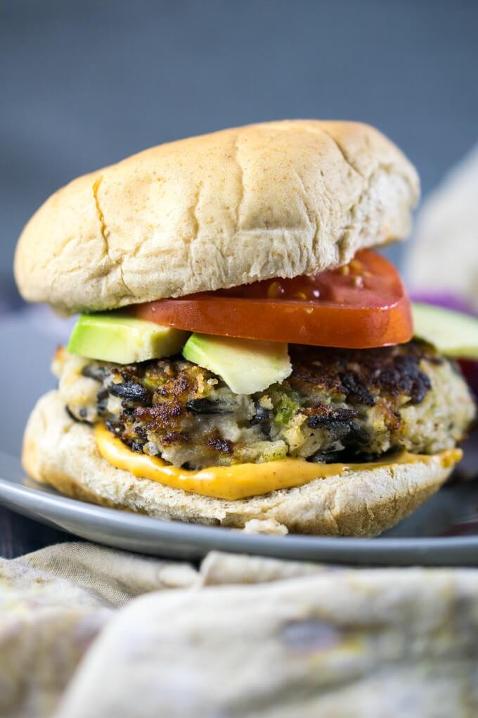 Wild rice burgers made from rice, celery, onion, and white beans, on a burger bun with chipotle sauce, tomato, and avocado, sitting on a gray plate.