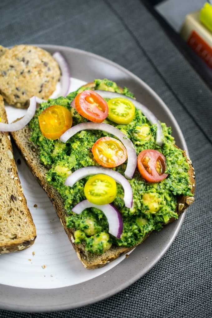 Whole grain toasted bread on a plate with crackers, with collard greens pesto and chickpeas spread on one slice and sliced cherry tomatoes and red onions arranged on top.