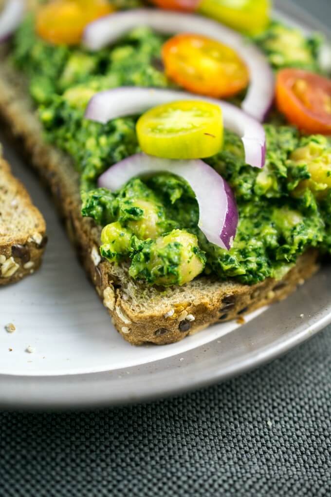 Close-up of garbanzo beans tossed in collard green pesto with grape tomatoes on a piece of seeded bread.