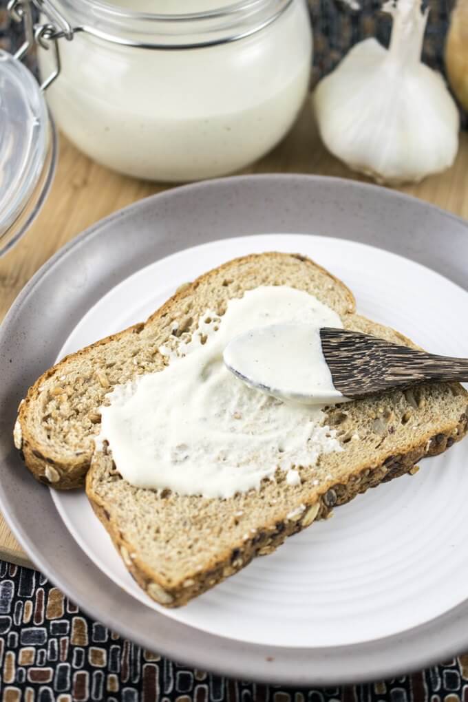 A piece of whole grain toast on a plate, with a spoonful of no-oil vegan mayonnaise being spread onto it, and a jar of mayo and bulb of garlic in the background