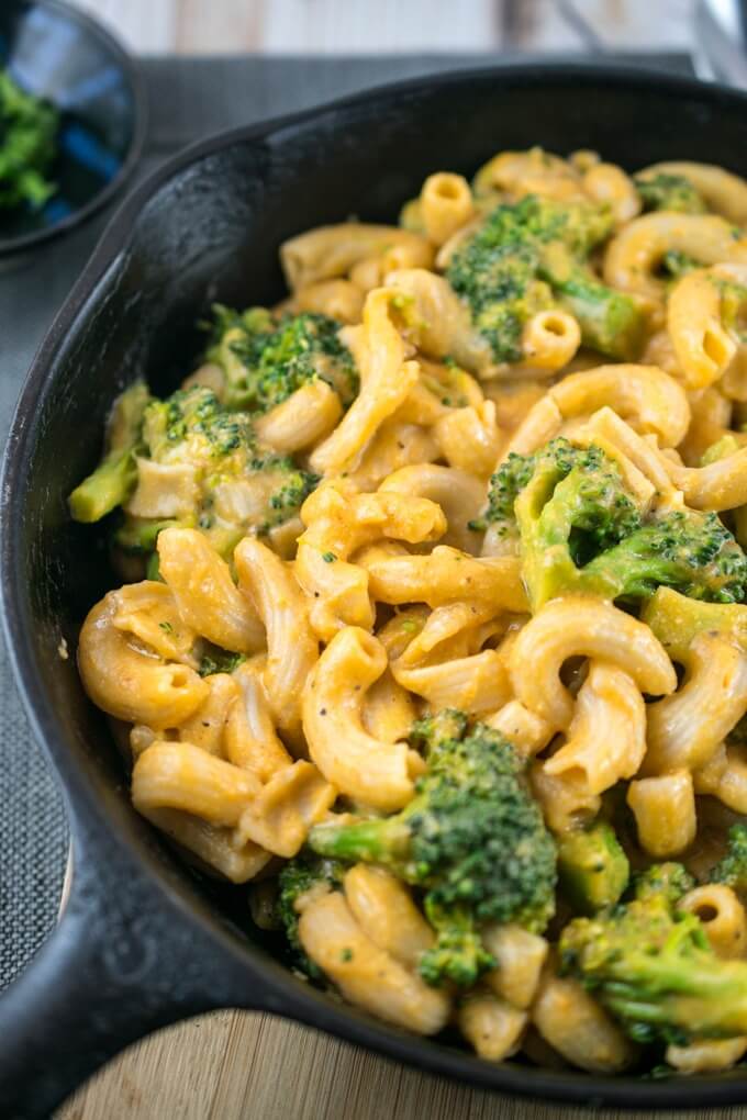 Broccoli and noodles in a skillet on a gray napkin with vegan cheese sauce and freshly-ground black pepper, and a bowl of basil in the background