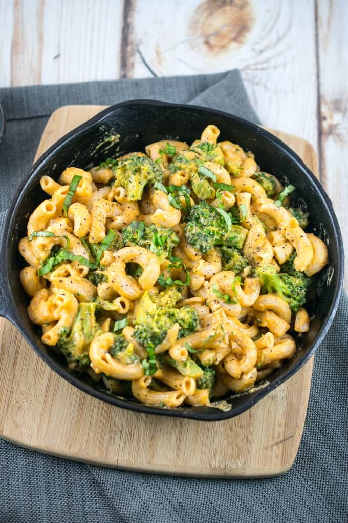 Vegan black pepper cheesy mac and broccoli in a cast iron skillet on a wooden cutting board with a wooden background, garnished with fresh basil and red pepper flakes.