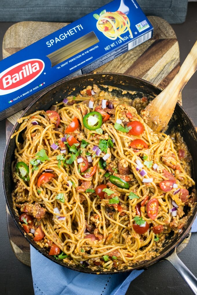 Overview view of a pan of cooked one pot taco pasta with a box of Barilla Spaghetti, cherry tomato slices, jalapeno slices, red onion and fresh coriander. Tempeh crumbles are visible on the pasta.