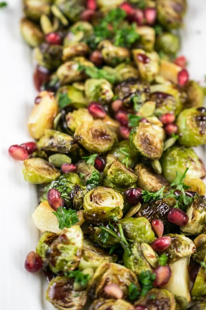 A pile of roasted Brussels sprouts on a white background with pomegranate seeds, pepitas, chopped parsley, and a dark purple pomegranate glaze
