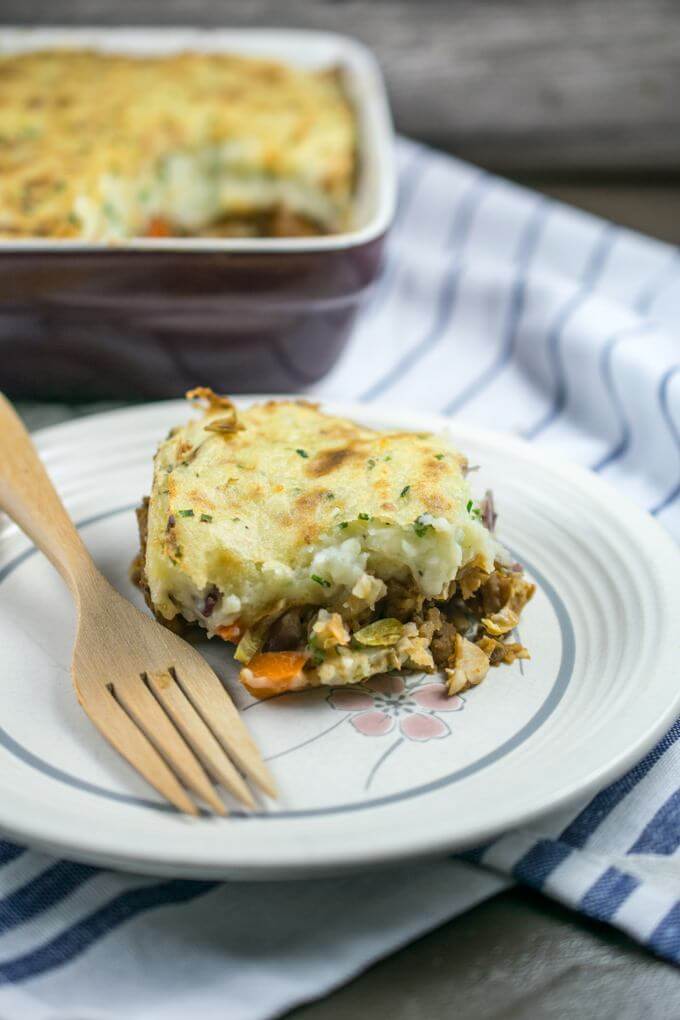 A slice of lentil vegan shepherd's pie on a plate with a wooden fork and the rest of the casserole in the background