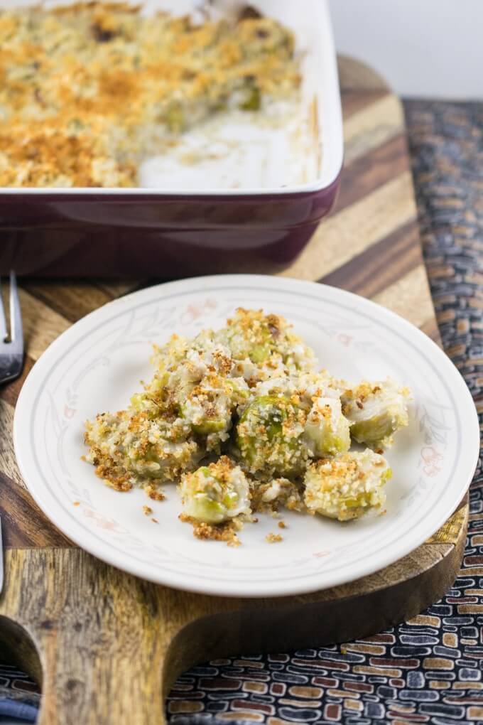 A serving of vegan Brussels sprout gratin on a plate atop a wooden cutting board next to the rest of the casserole in a baking dish