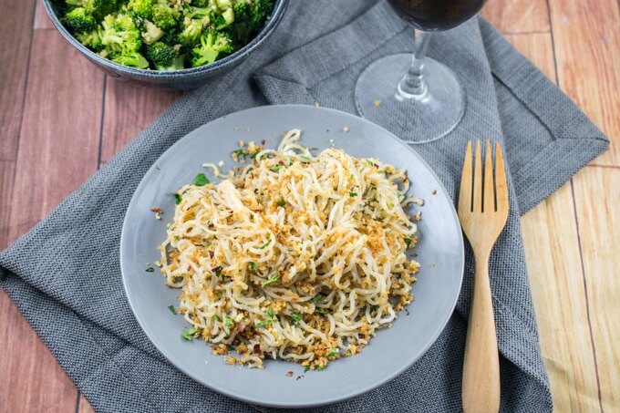 Overhead view of a table setting with a gray napkin, glass of wine, spaghetti with breadcrumbs and fresh parsley, a wooden fork, and a side of broccoli.