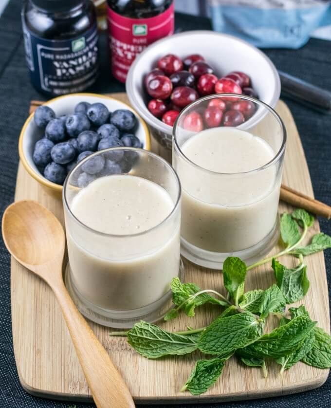 Two ramekins of vegan vanilla pudding arranged on a cutting board with bowls of blueberries and cranberries, a wooden spoon, cinnamon sticks, fresh mint, and bottles of vanilla extract and peppermint extract in the background.