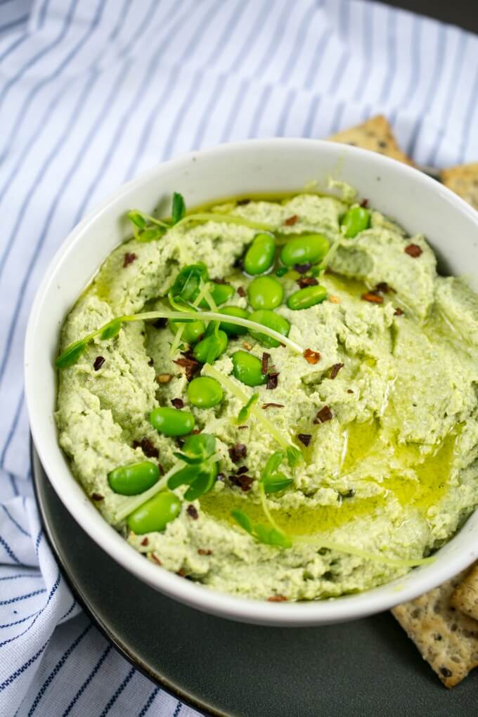 A bowl of bright green edamame hummus drizzled with olive oil and garnished with fresh edamame, broccoli sprouts, and red pepper flakes