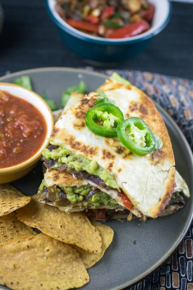 A stack of three vegan quesadillas on a plate, with layers of avocado and cooked vegetables visible, and a small bowl of salsa with tortilla chips.
